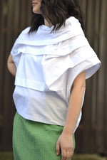 Bluză albă cu design asimetric și falduri decorative suprapuse, fotografiată din lateral.