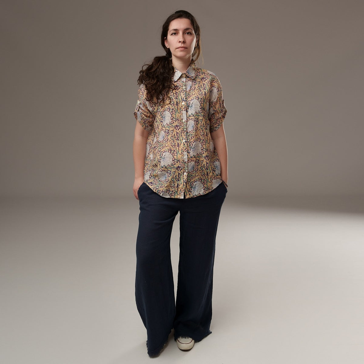 Cămașă din rayon cu imprimeu floral în nuanțe calde de bej, mânecă scurtă și croială lejeră, purtată cu pantaloni largi bleumarin.