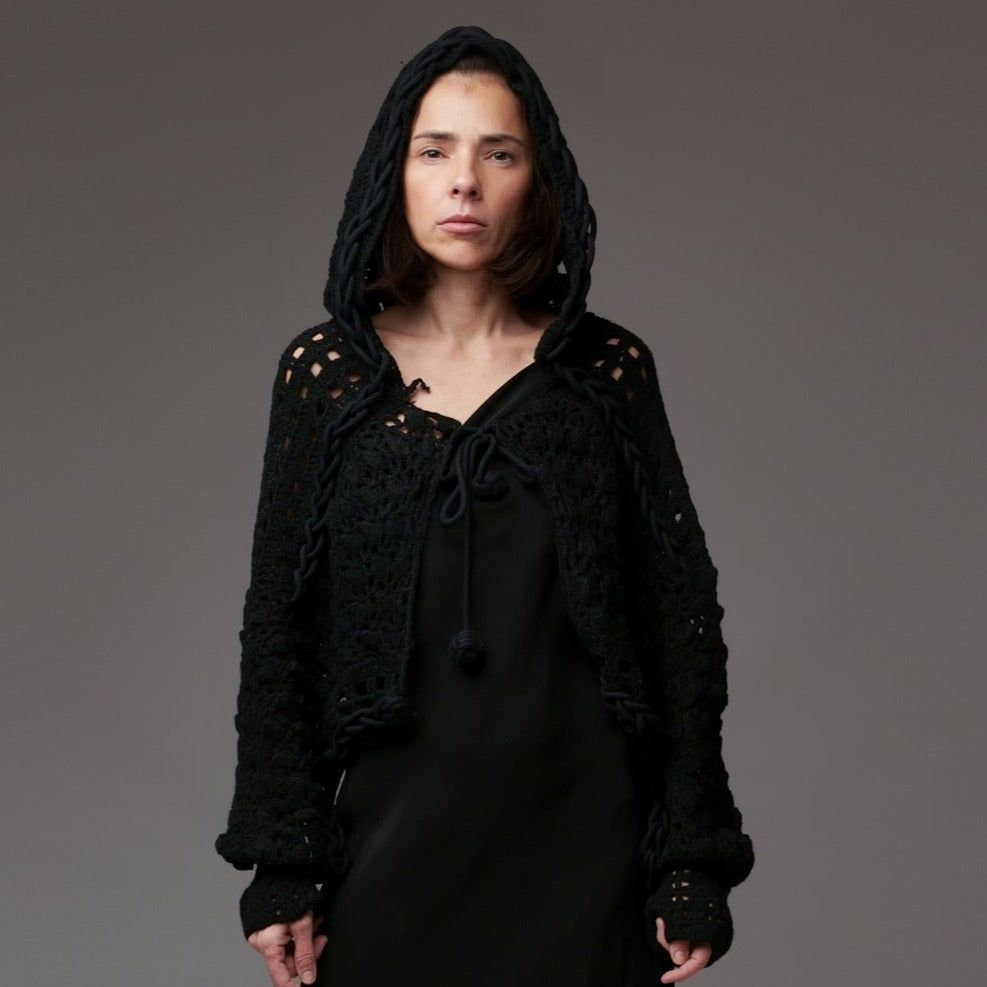 Capă stil bolero croșetată din acril negru, cu glugă supradimensionată și șireturi groase decorative, purtată peste rochie neagră.
