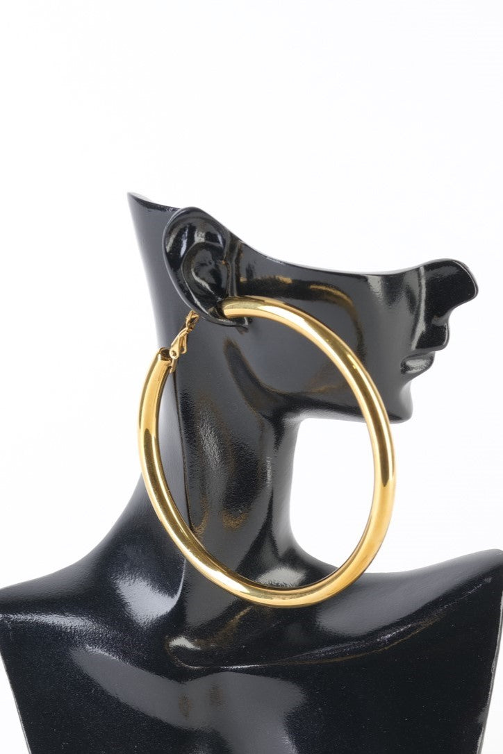 Pereche de cercei aurii tip hoop din oțel inoxidabil, diametru 7 cm, unul purtat pe manechin și celălalt așezat pe fundal alb.