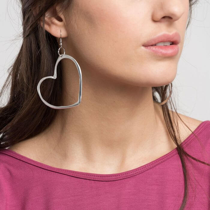 Cercei cu pandantiv în formă de inimă, purtați de o femeie cu părul deschis la culoare, zâmbind și având un look casual cu un top roz.