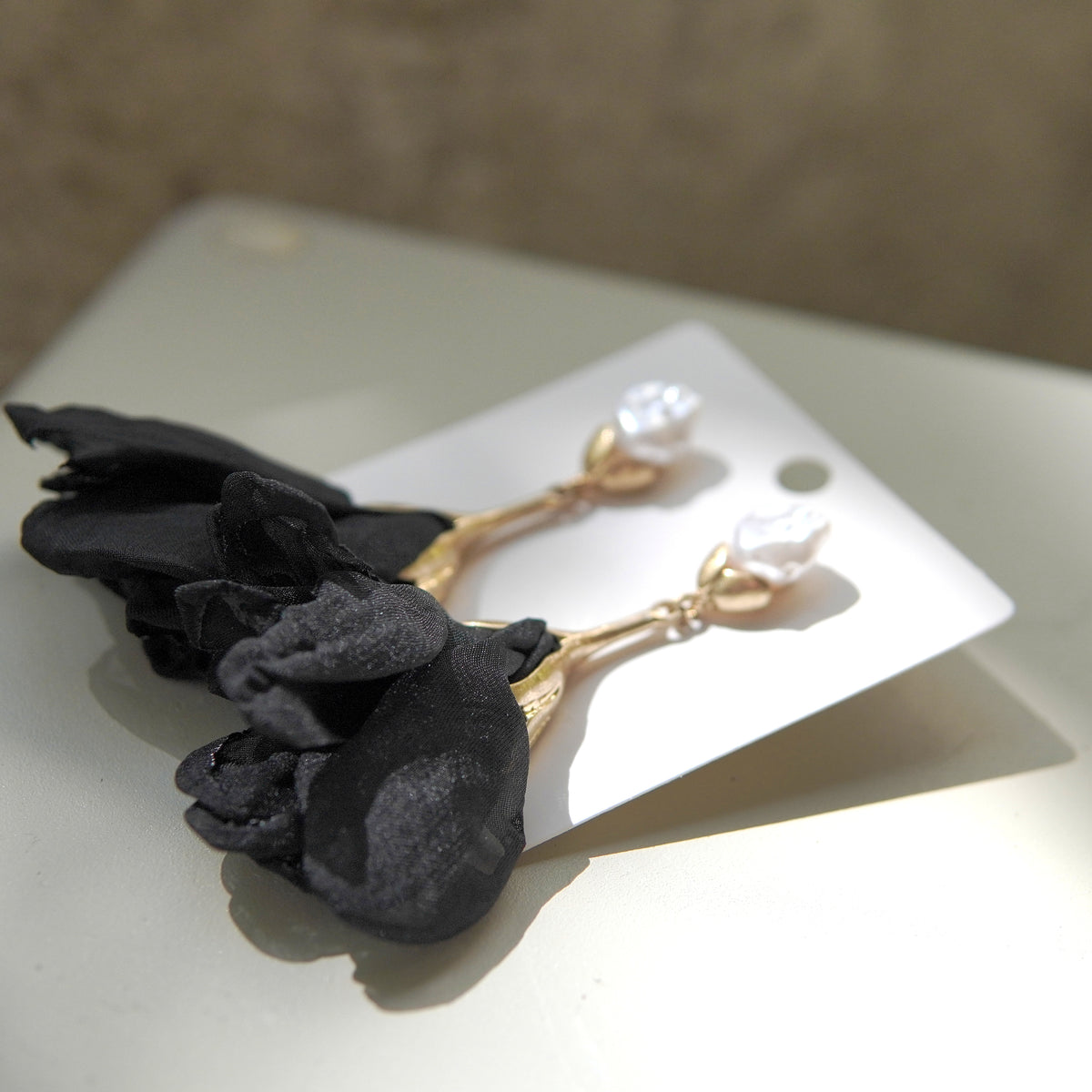 Pereche de cercei din oțel inoxidabil cu perlă și floare textilă neagră, prezentați pe suport alb.