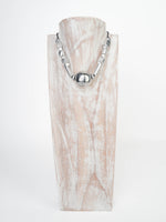 Colier ajustabil Vestopazzo din aluminiu reciclat, cu design geometric și detaliu central rotund, expus pe suport de lemn.