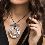 Colier cu pandantiv format din cercuri concentrice din aluminiu reciclat, purtat la gât pe șnur negru reglabil.