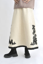 Fustă lungă evazată din tricot crem, cu broderie neagră pe tiv, purtată cu pulover supradimensionat.