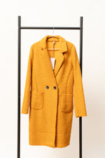 Palton lung damă în nuanță muștar, confecționat din stofă boucle, cu croială clasică și buzunare mari.