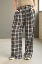 Pantaloni drepți din stofă în carouri maro-bej, purtați cu talia evidențiată de o curea subțire maro.