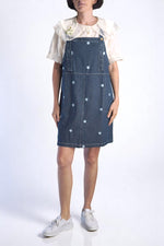 Rochie tip salopetă din denim bleumarin cu broderii florale albastre, purtată cu cămașă crem din bumbac.