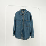 Jachetă tip cămașă din denim albastru, cu buzunare mari și închidere cu capse, expusă pe un umeraș alb.