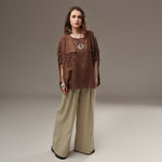 luză lejeră maro din in cu mâneci tip fluture, ușor bufante și încrețite lateral, purtată cu pantaloni largi crem. Modelul poartă colier geometric și brățări metalice.

