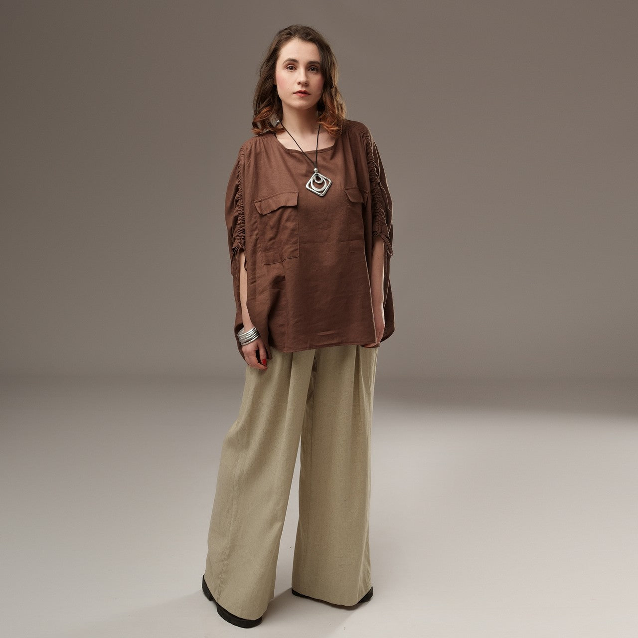 luză lejeră maro din in cu mâneci tip fluture, ușor bufante și încrețite lateral, purtată cu pantaloni largi crem. Modelul poartă colier geometric și brățări metalice.

