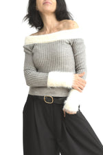 Bluză tip pulover din tricot gri, cu decolteu lăsat pe umeri și manșete din fir pufos alb, purtată cu pantaloni negri.