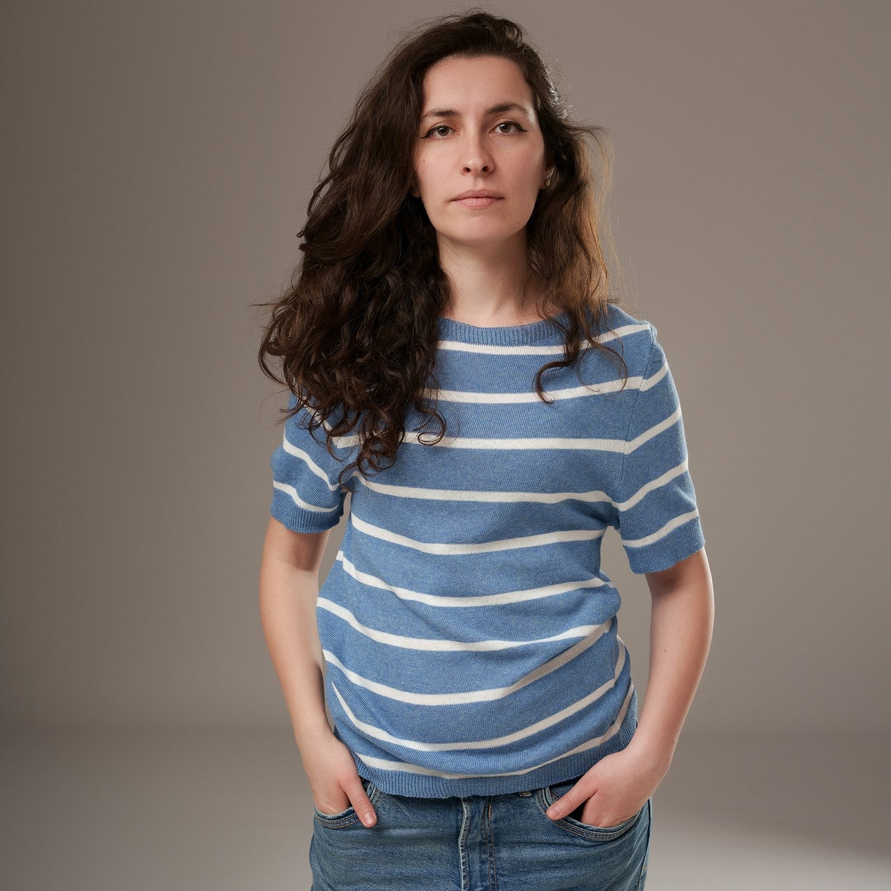 Bluză din tricot subțire bleumarin cu dungi albe, mânecă scurtă și guler rotund, purtată de model cu mâinile în buzunar.