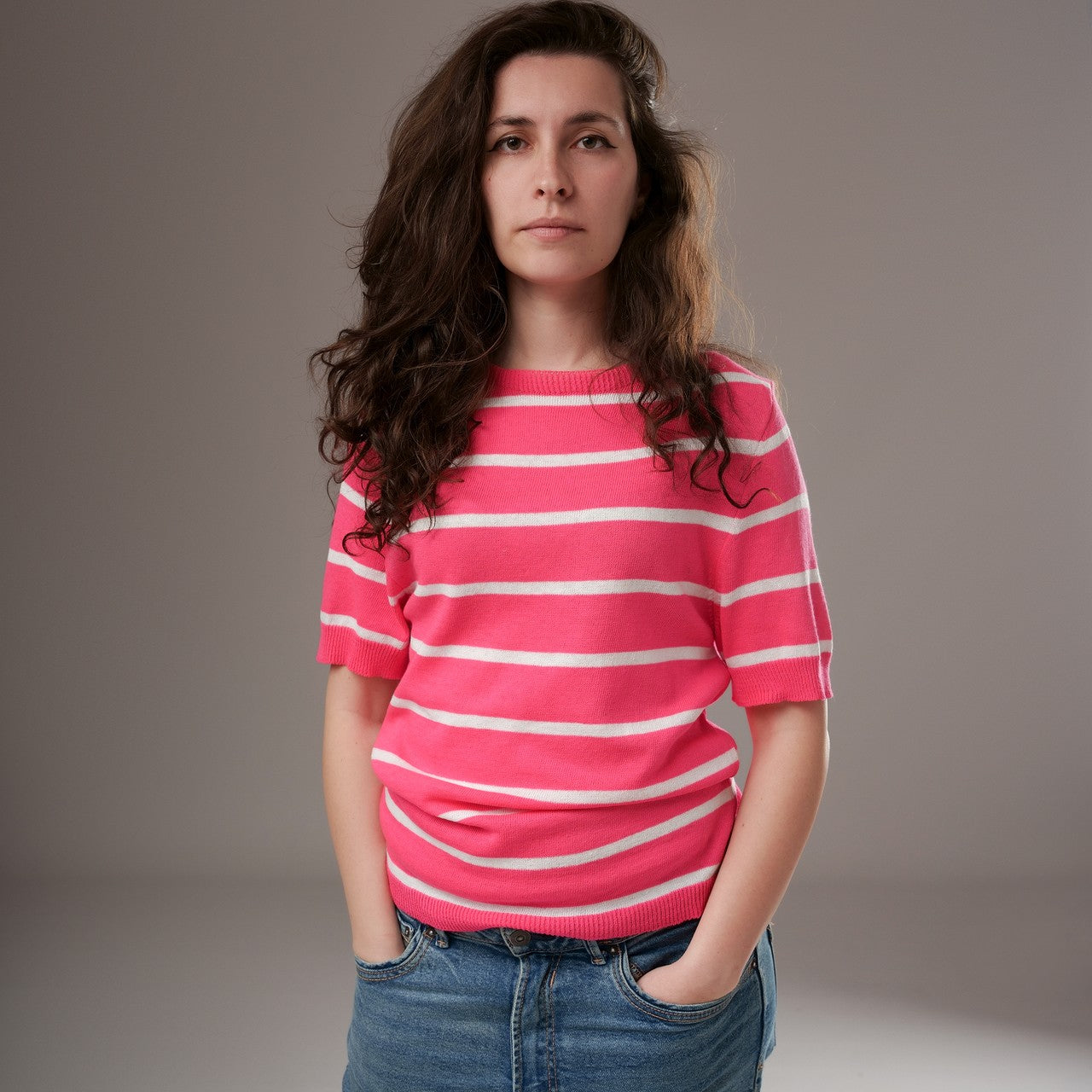 Bluză casual roz cu dungi albe orizontale, din tricot subțire, cu guler rotund și mânecă scurtă, purtată de model cu mâinile în buzunar.