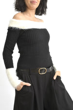 Vedere laterală a bluzei negre din tricot, cu umeri goi și mâneci lungi terminate cu manșete pufoase albe.