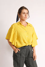 Cămașă galbenă din in purtată legată în față tip crop top, cu mâneci fluture și închidere cu nasturi.
