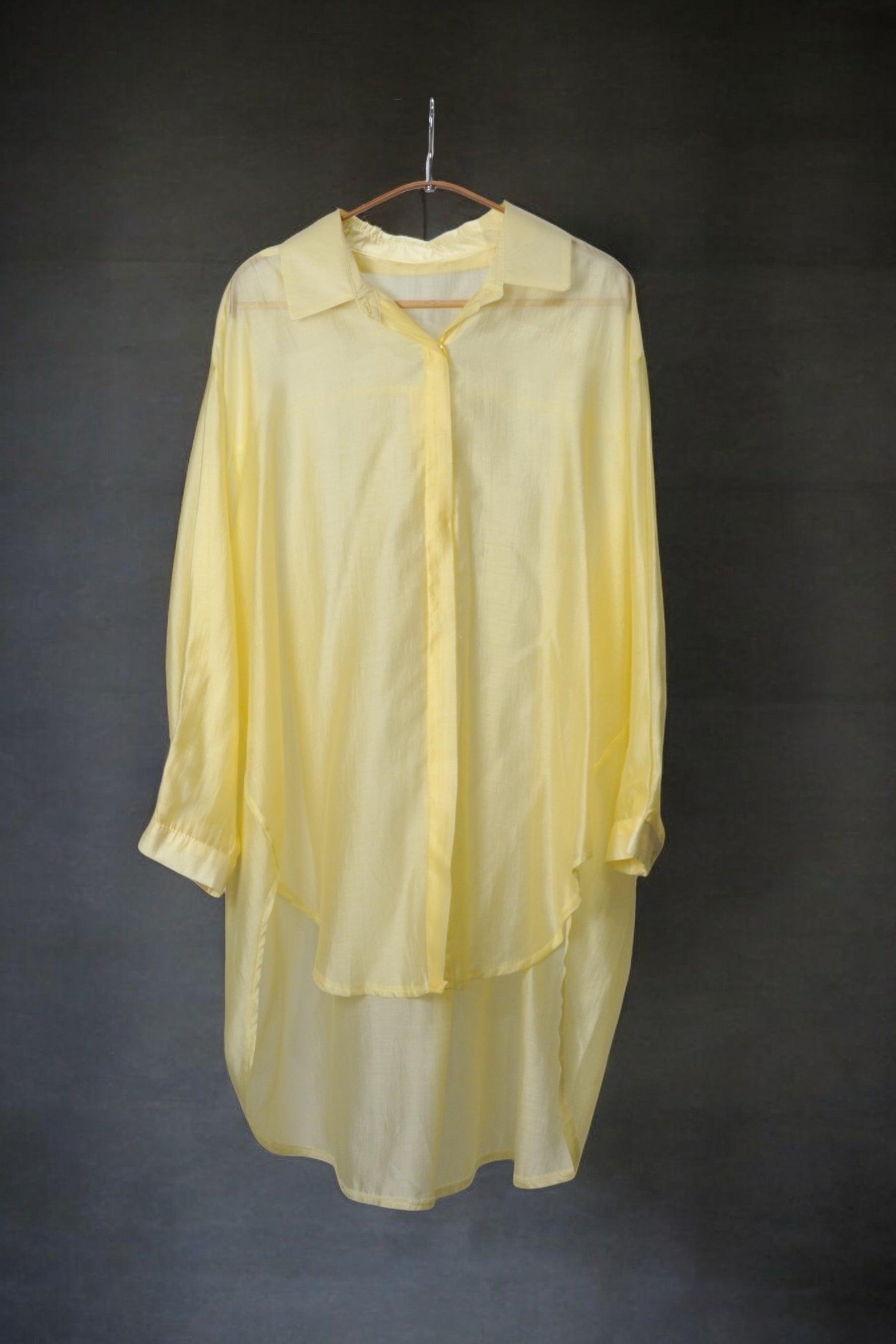 Cămașă oversized galben pastel, cu textură fină și material vaporos, pe umeraș, fundal închis.
