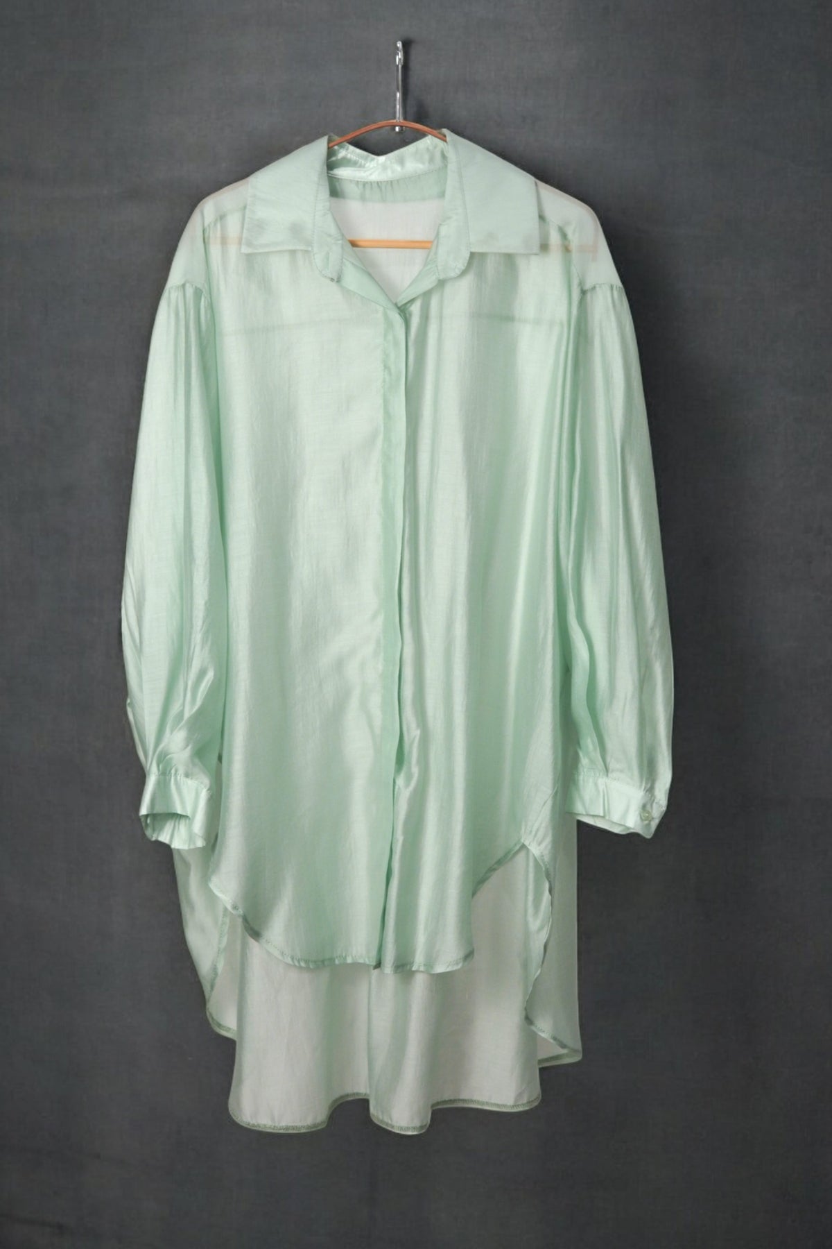 Cămașă oversized verde pastel, cu aspect lejer și croială fluidă, pe umeraș, fundal închis.