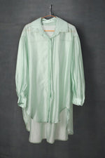 Cămașă oversized verde pastel, cu aspect lejer și croială fluidă, pe umeraș, fundal închis.