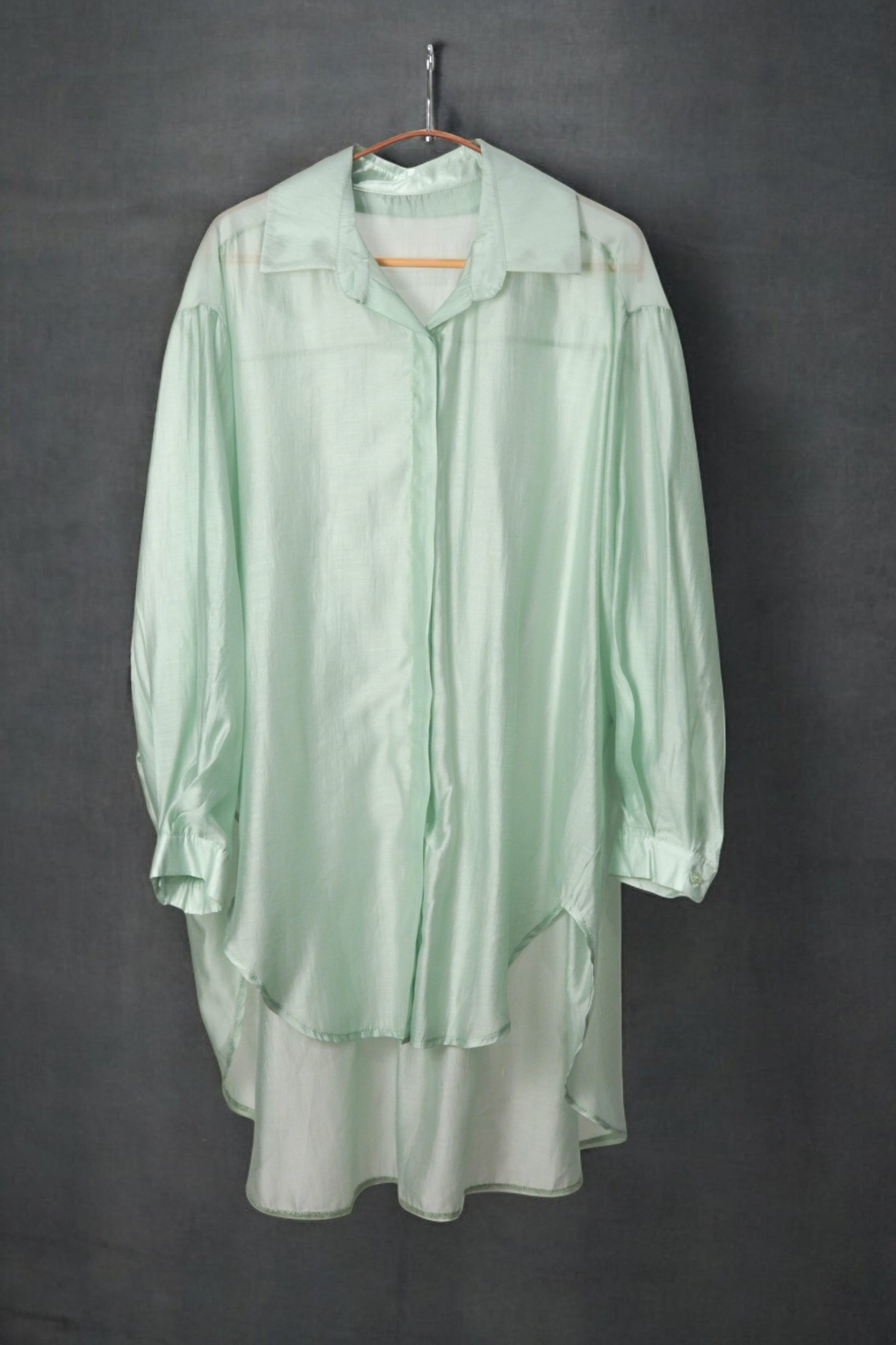 Cămașă oversized verde pastel, cu aspect lejer și croială fluidă, pe umeraș, fundal închis.