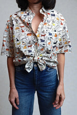 Cămașă cu mânecă scurtă, imprimeu cu pisici naive și închidere cu nod frontal, purtată cu jeanși.