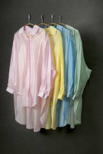 Set de cămăși oversized în culori pastel – roz, galben, bleu și verde, aranjate pe umerașe, pe fundal negru.