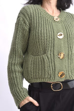 Detaliu cardigan tricotat verde cu ochiuri mari și nasturi aurii supradimensionați.
