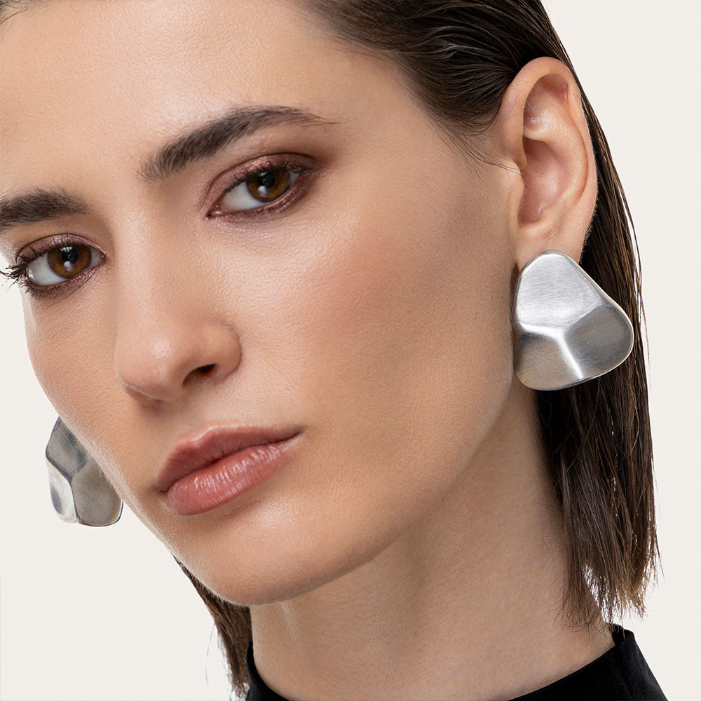 Cercei în formă de piatră din aluminiu reciclat, cu design organic și texturat, purtați de o femeie pentru un look sofisticat.