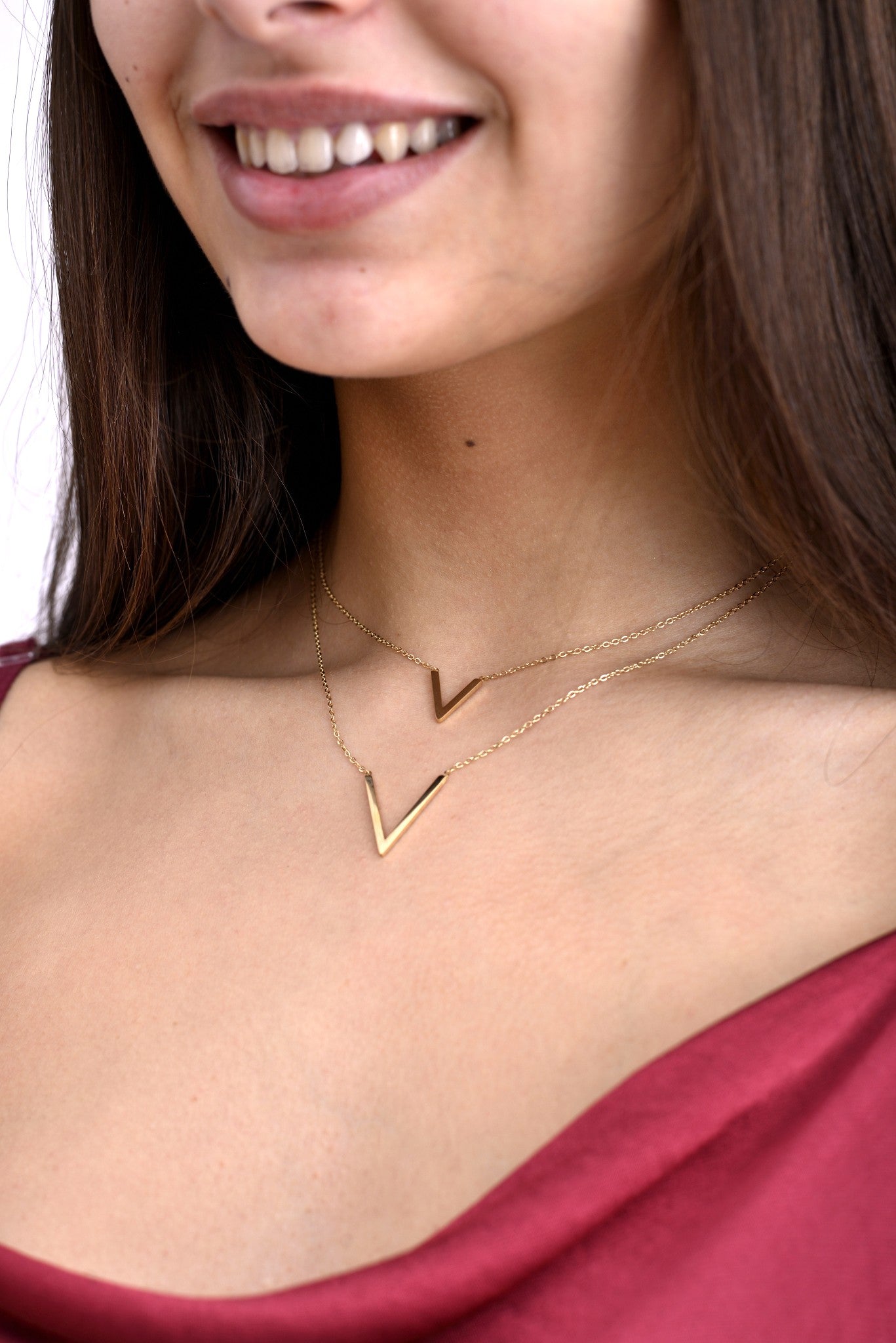Detaliu apropiat al unui colier dublu auriu cu pandantive în formă de V, purtat la un top satinat bordo.