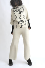 Compleu bej format din top cu imprimeu grafic tip portret asimetric și pantaloni largi, purtat cu adidași negri.