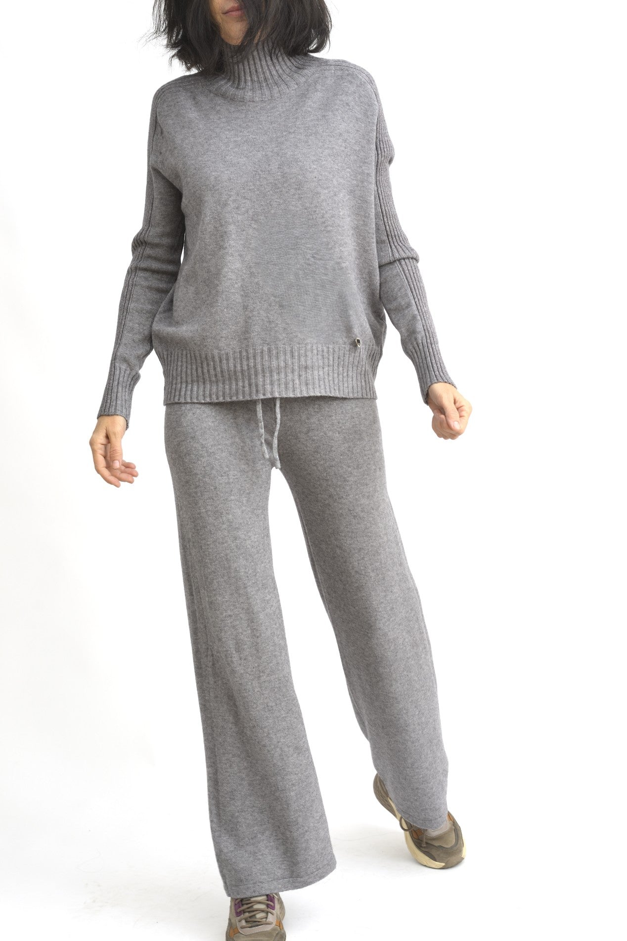 Vedere dinamica a compleului gri din tricot, format din top căzut pe umeri și pantaloni largi.