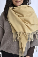 Eșarfă bej deschis, cu franjuri fini, surprinsă în detaliu pentru a evidenția textura moale și căderea naturală a materialului.