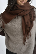 Eșarfă maro, cu franjuri discrete, așezată în jurul gâtului peste un pulover bej, într-o stilizare calmă și versatilă.