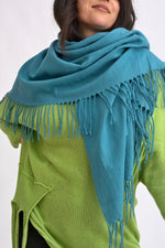 Eșarfă turcoaz cu franjuri fine, purtată lejer peste un pulover verde, evidențiind textura moale și drapajul natural.