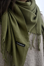Detaliu de eșarfă verde măsliniu cu franjuri și etichetă „Cashmere”, evidențiind finisajul și textura plăcută a materialului.