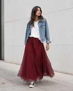 Model poartă fustă lungă din tul burgundy cu bandă satinată, tricou alb lejer, jachetă scurtă din denim și teniși Converse.