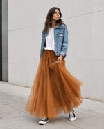 Model poartă fustă lungă din tul cărămiziu cu bandă satinată, tricou alb lejer, jachetă scurtă din denim și teniși Converse.