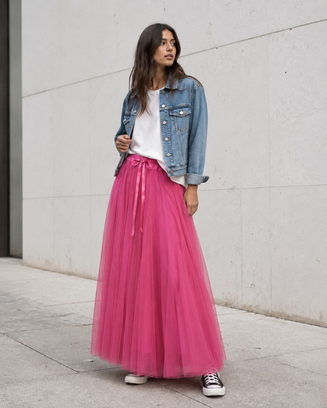 Model poartă fustă lungă din tul roz intens cu bandă satinată, tricou alb lejer, jachetă scurtă din denim și teniși Converse.
