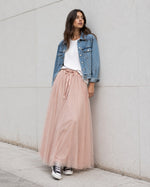 Model poartă fustă lungă din tul roz pudrat cu bandă satinată, tricou alb lejer, jachetă scurtă din denim și teniși Converse.