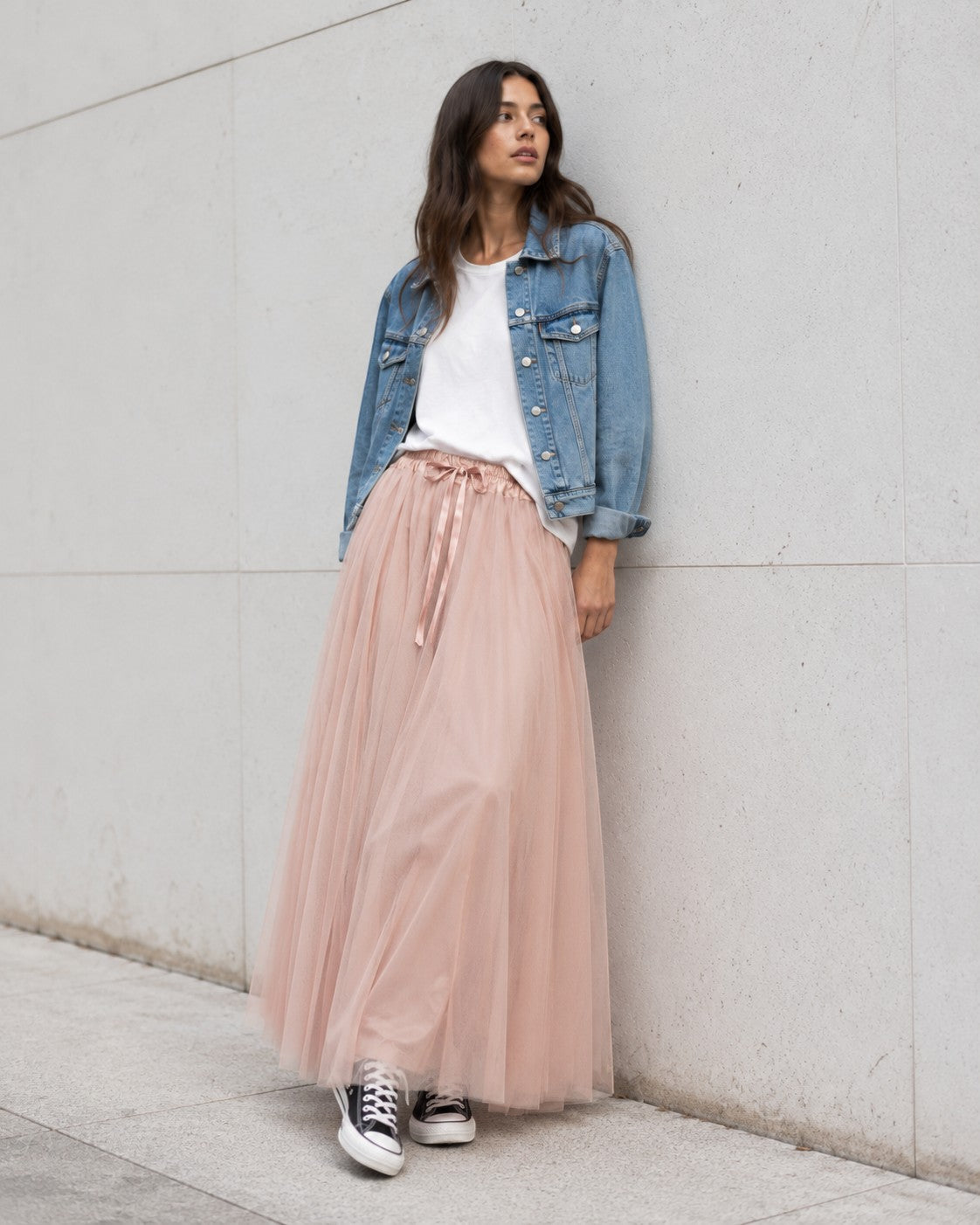 Model poartă fustă lungă din tul roz pudrat cu bandă satinată, tricou alb lejer, jachetă scurtă din denim și teniși Converse.