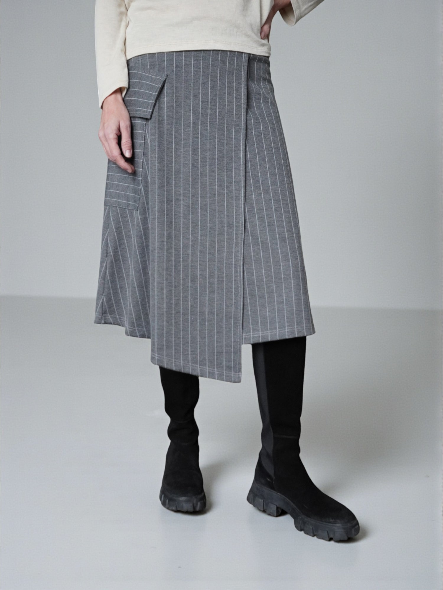 Fustă midi în dungi fine, cu colț suprapus și croială asimetrică, purtată cu cizme negre.