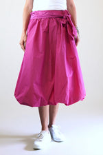 Fustă fucsia cu croială gogoșar și fundă laterală decorativă, realizată din material ușor și lucios, stil casual-modern