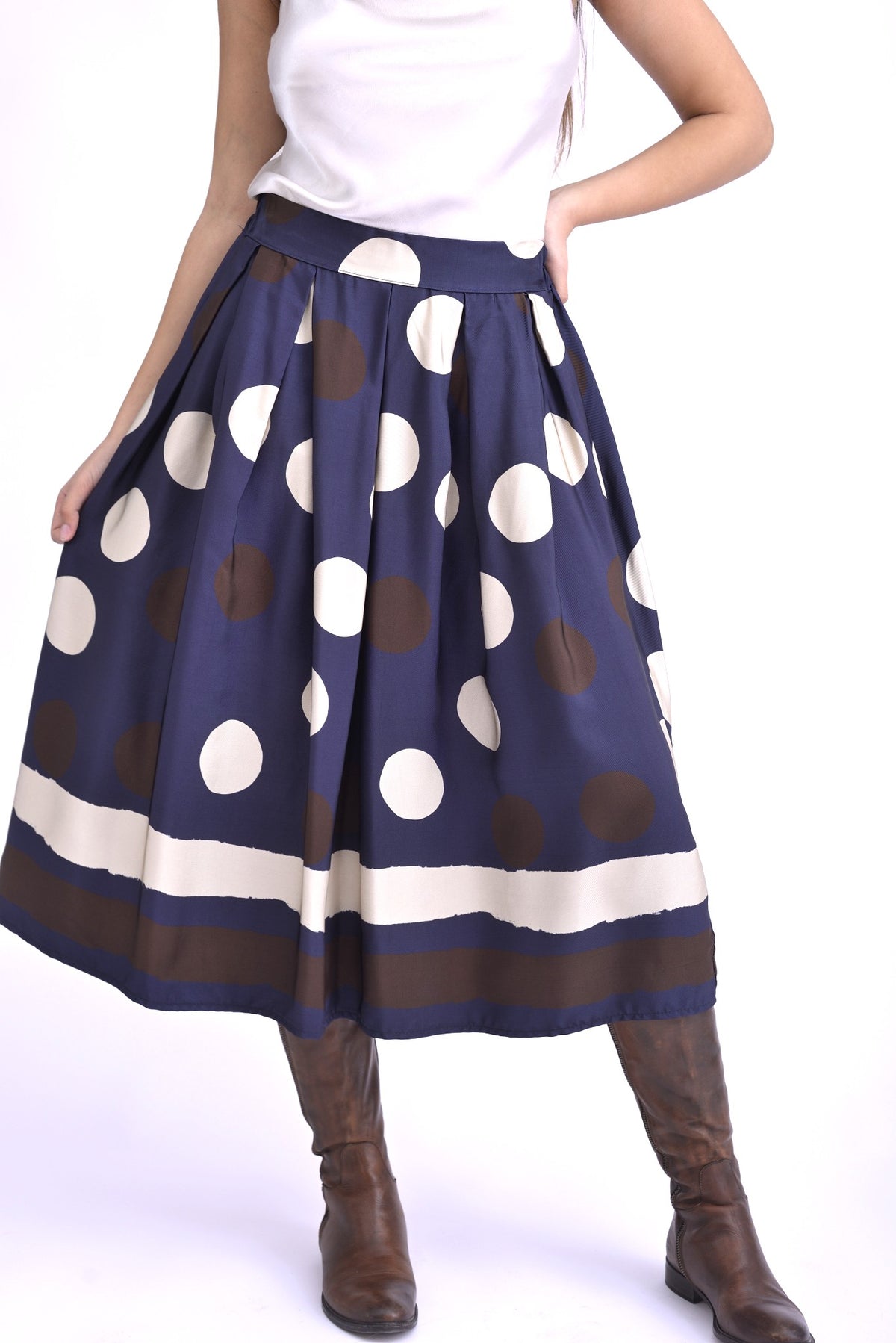 Model purtând o fustă midi bleumarin cu imprimeu cu buline mari maro și albe, completată de cizme maro și top satinat deschis.