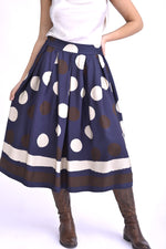 Model purtând o fustă midi bleumarin cu imprimeu cu buline mari maro și albe, completată de cizme maro și top satinat deschis.