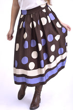 Model purtând o fustă midi bleumarin cu buline albe și maro, stilizată cu top satinat gri închis și cizme maro, pentru un look elegant de zi.