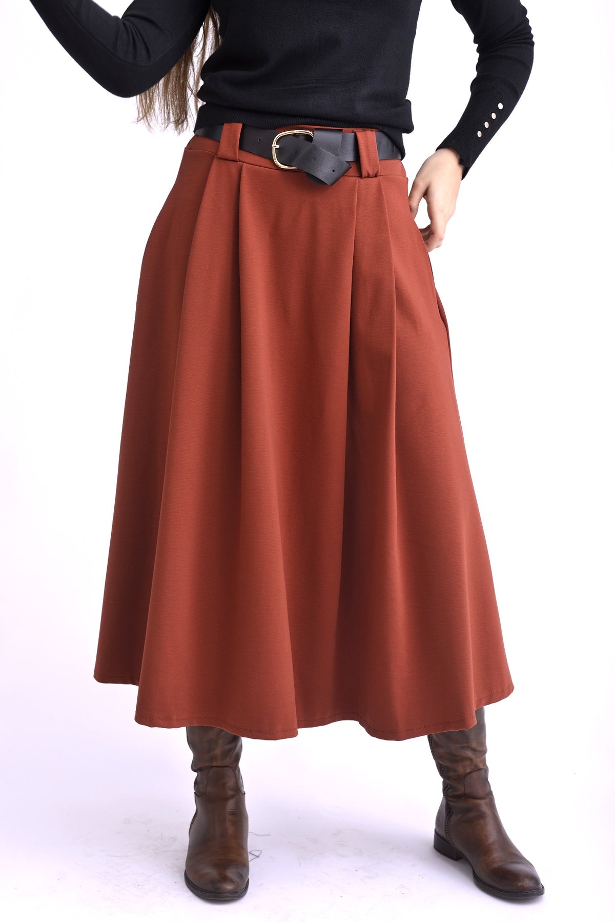 Model purtând o fustă midi cărămizie cu pliuri largi și curea neagră, asortată cu pulover negru și cizme maro.