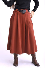 Model purtând o fustă midi cărămizie cu pliuri largi și curea neagră, asortată cu pulover negru și cizme maro.