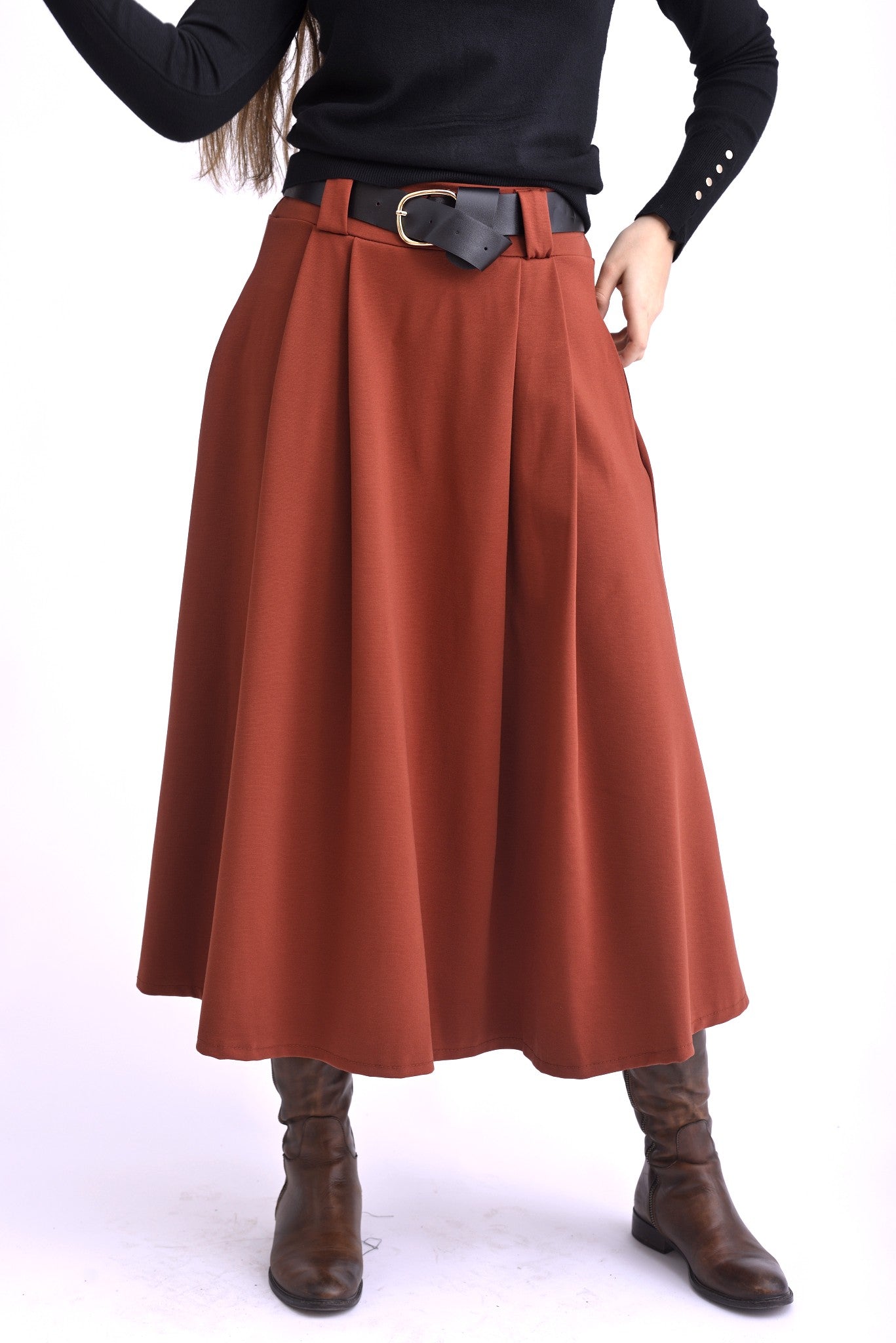 Model purtând o fustă midi cărămizie cu pliuri largi și curea neagră, asortată cu pulover negru și cizme maro.