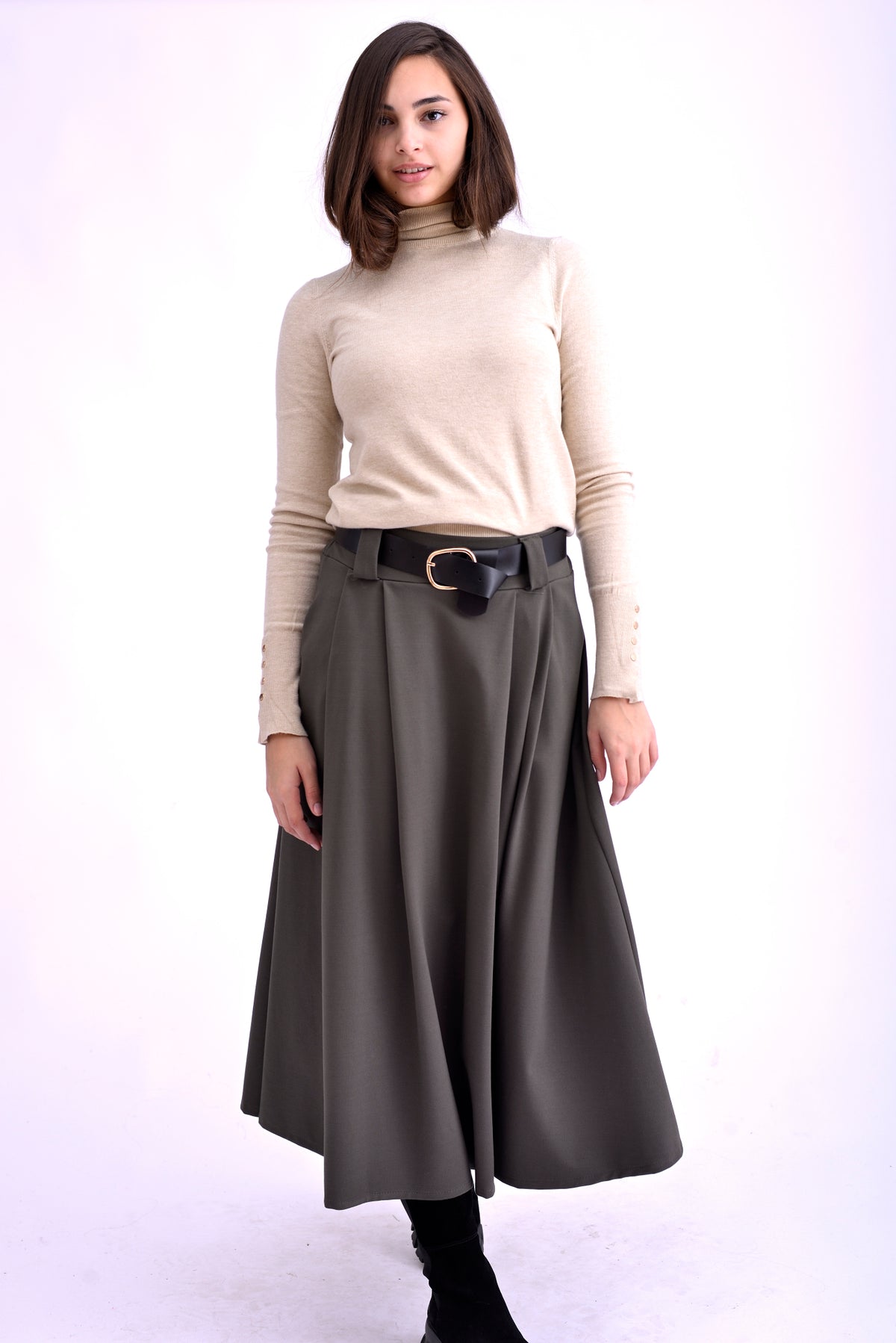 Model purtând o fustă midi gri kaki cu pliuri, curea neagră cu cataramă aurie și pulover bej cu guler înalt, asortată cu ghete negre.
