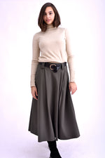 Model purtând o fustă midi gri kaki cu pliuri, curea neagră cu cataramă aurie și pulover bej cu guler înalt, asortată cu ghete negre.